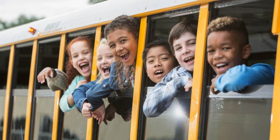 A group of six multi-ethnic elementary school students, 7 to 9 years old, on a yellow school bus, looking out the windows. The view is from outside the bus looking in.