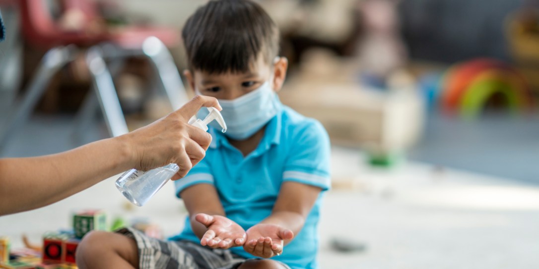 Young asian boy wearing a protective face mask sanitizes his hands while playing at daycare.
